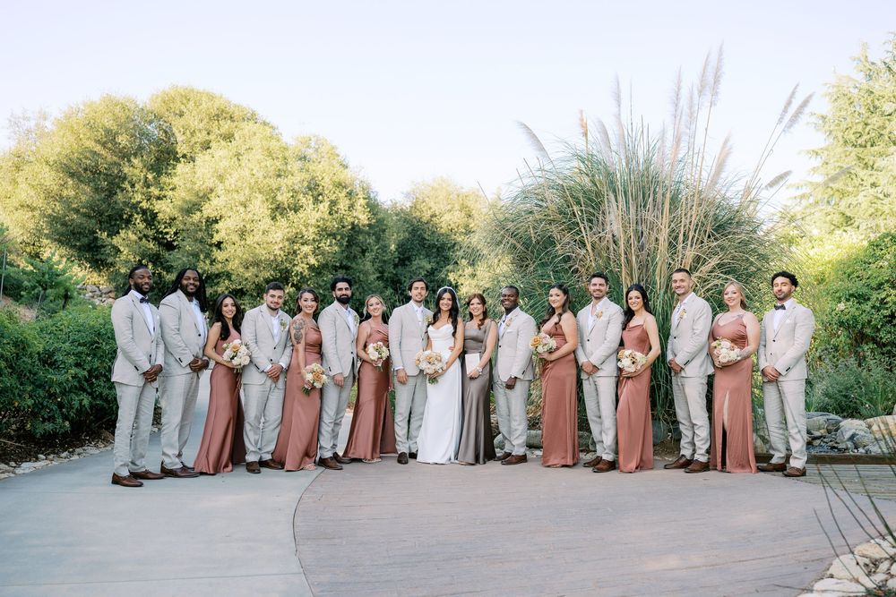Wedding party poses together outdoors, bridesmaids in rust dresses and groomsmen in tan suits with lush greenery.