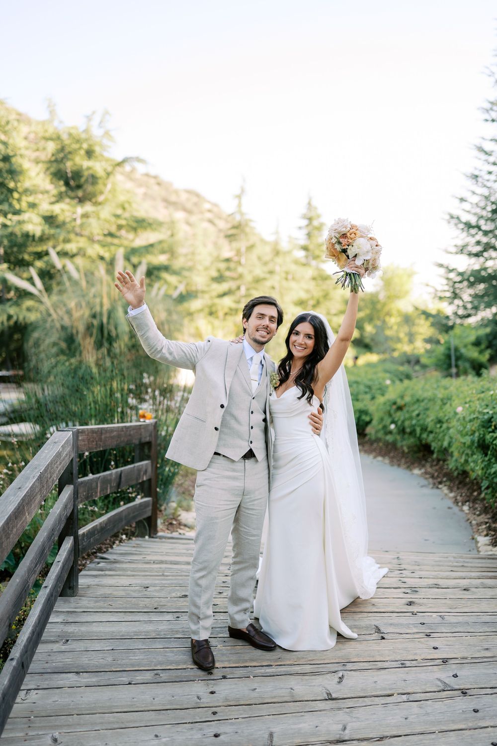 Happy newlywed couple celebrating on a wooden bridge, bride in white gown holding bouquet, groom in beige suit, lush greenery.
