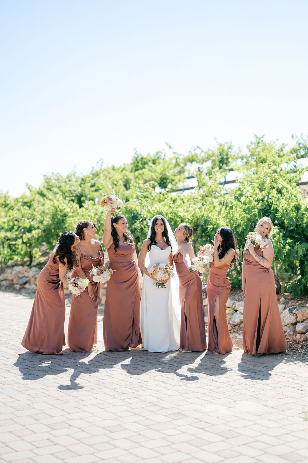 Bride in white gown poses with six bridesmaids in rust-colored satin dresses holding bouquets at a vineyard wedding.