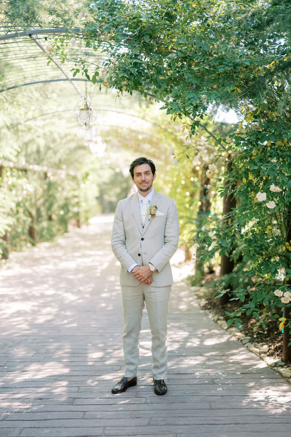 Elegant groom in beige three-piece suit standing on wooden path under lush green garden archway on wedding day.