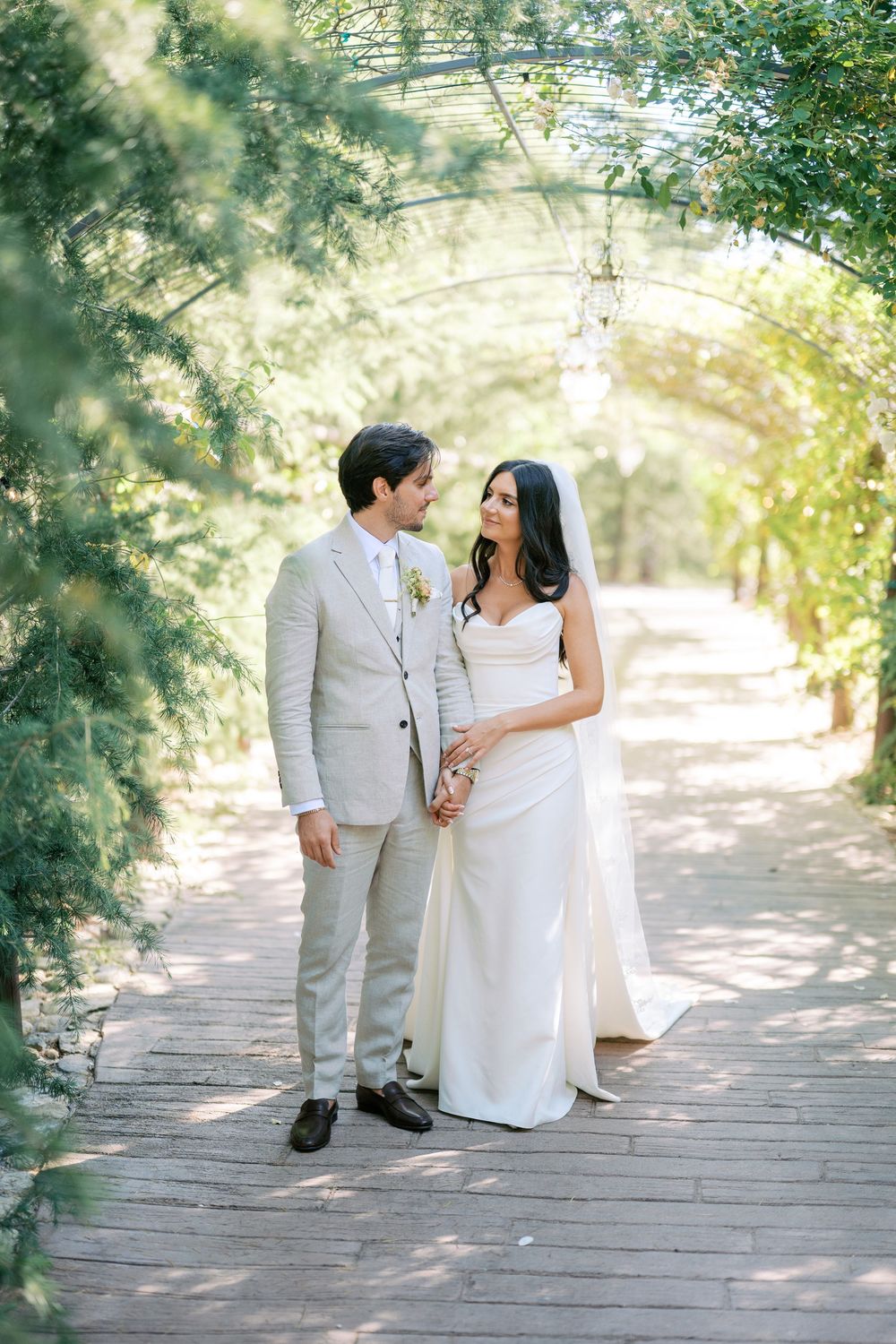Bride and groom share a romantic moment under a green floral arch on a wooden pathway at their wedding.
