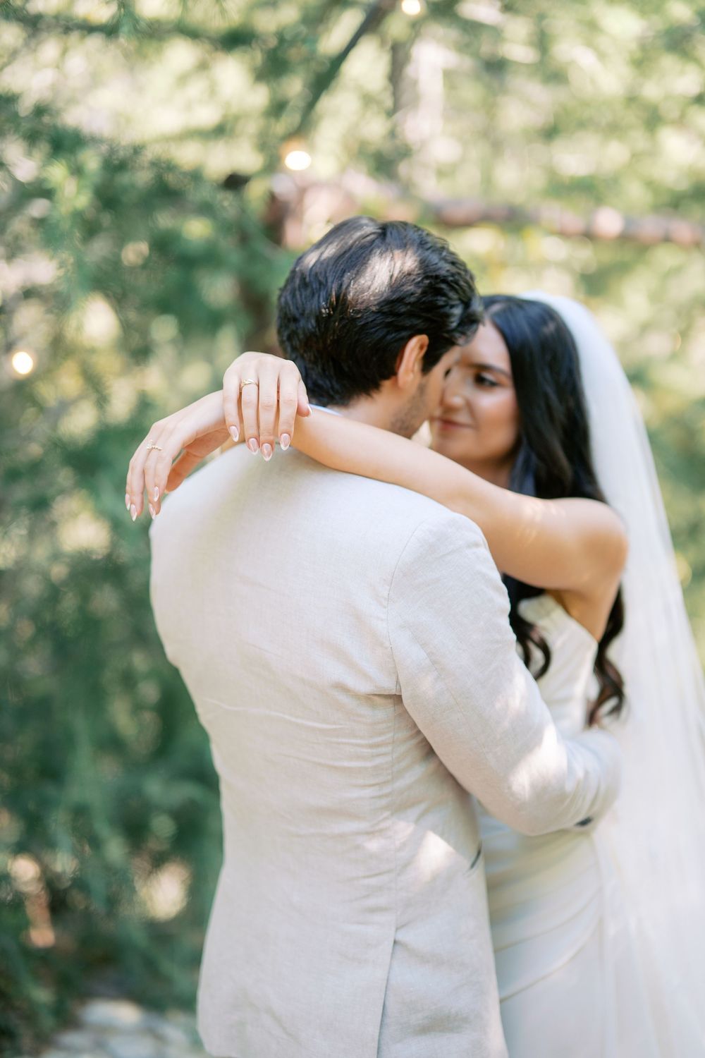 Bride in white veil embraces groom in beige suit outdoors, showcasing her engagement ring in a romantic moment.