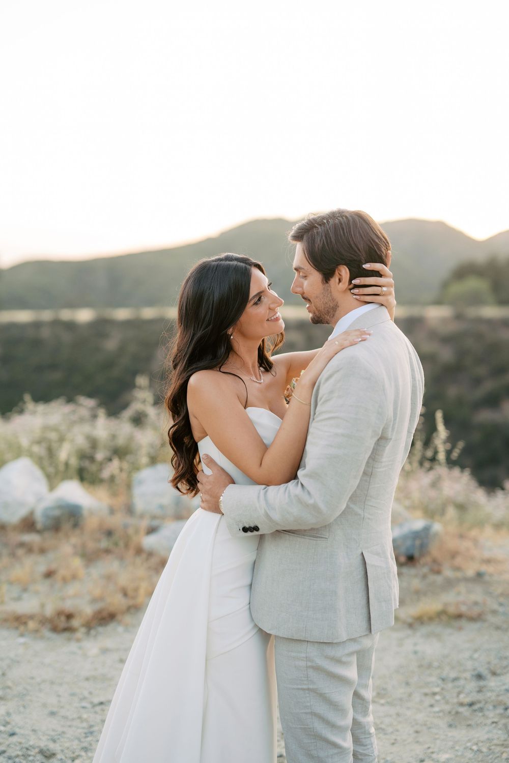 A bride and groom share a romantic embrace at golden hour with scenic mountains in the background.