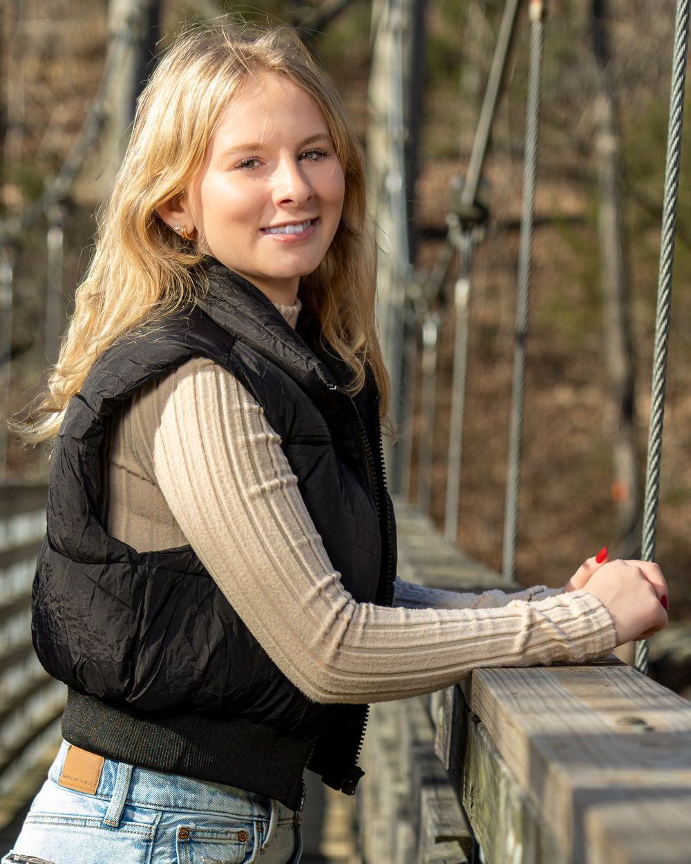 Young blonde woman smiling on a suspension bridge, wearing a black puffer vest and beige long-sleeve top.
