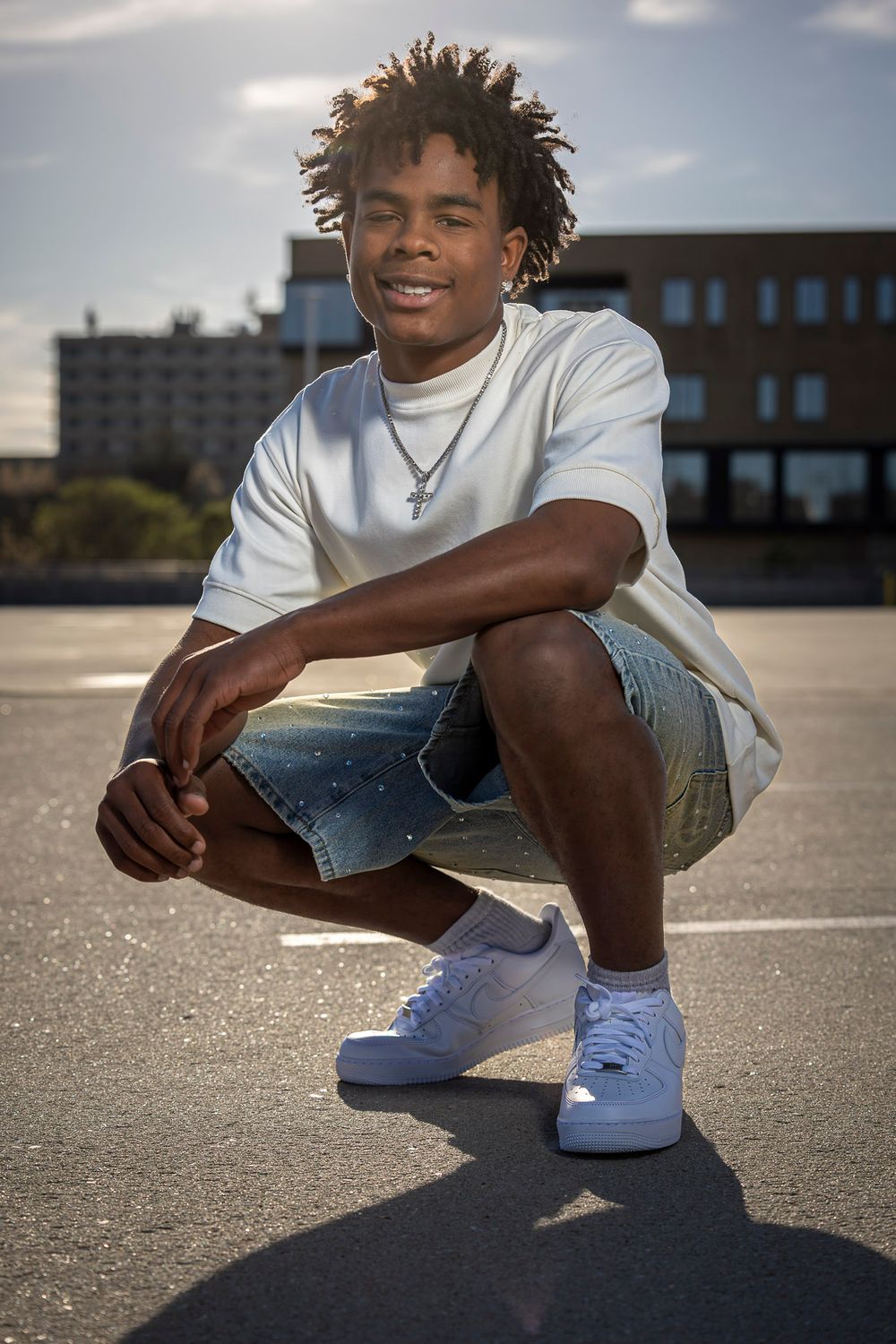 Young man crouching in parking lot wearing white tee, denim shorts, and white sneakers, smiling confidently.