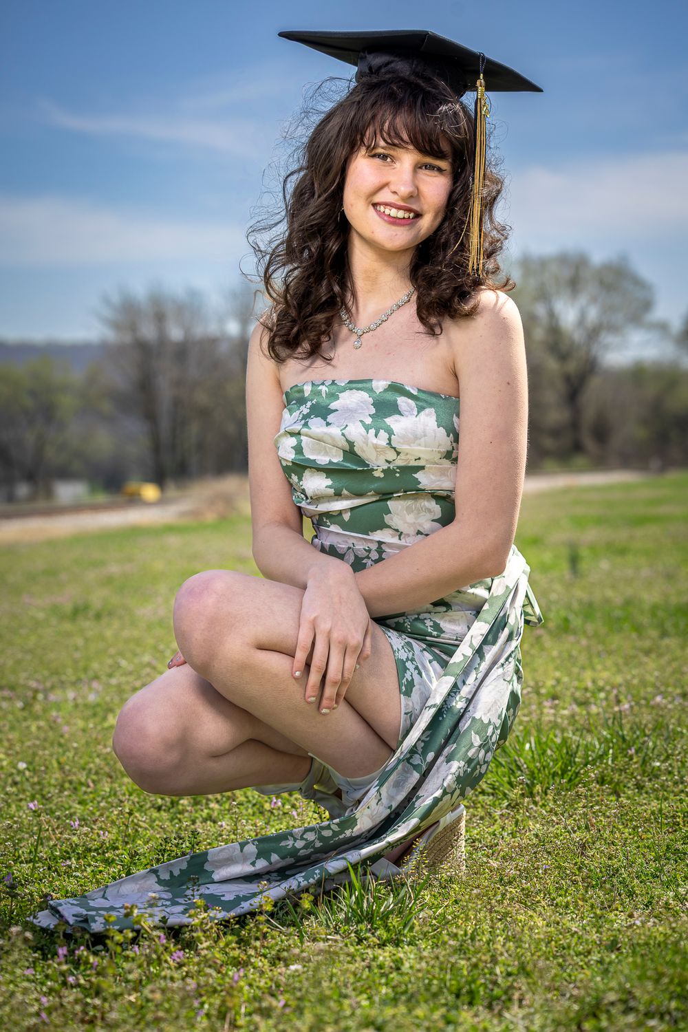 Smiling young woman in floral dress and graduation cap kneeling on green grass outdoors on a sunny day.