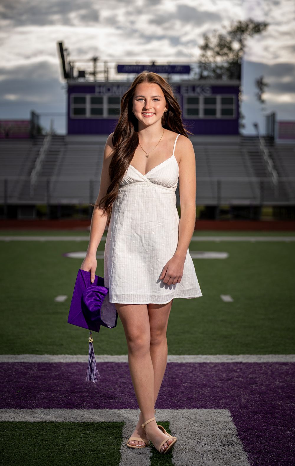 Young woman in white dress holding purple graduation cap on football field with purple stadium in background.