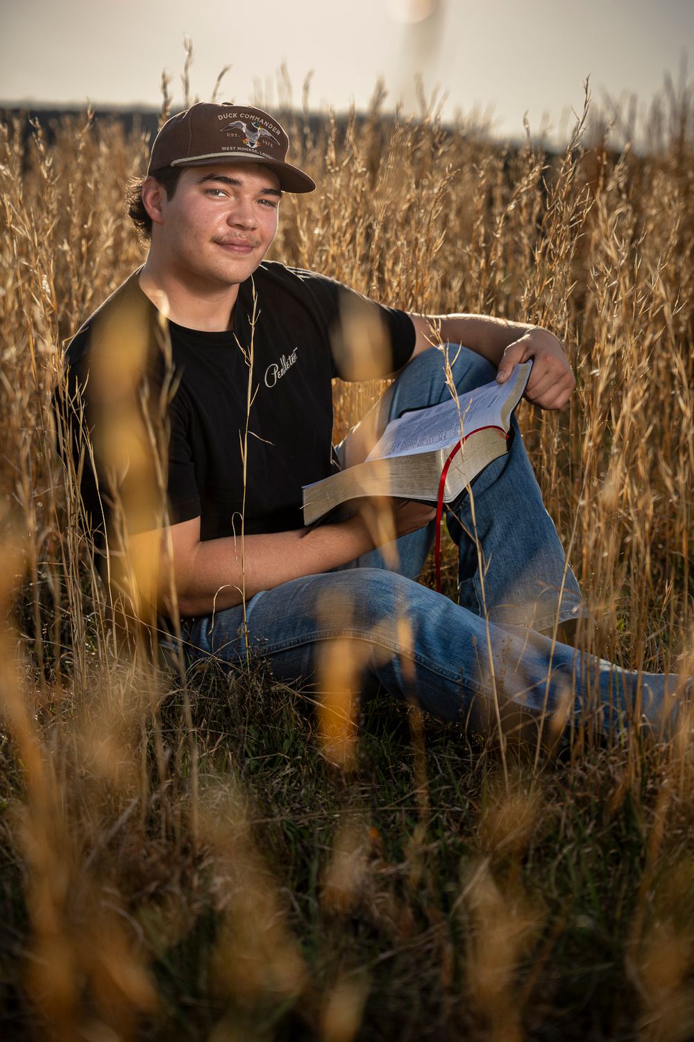 Young man in black cap and shirt sits in golden wheat field holding an open book with red ribbon bookmark.