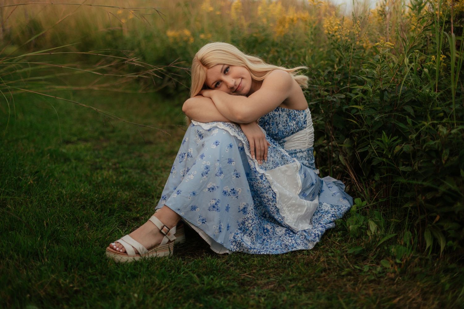 A senior girl sitting in the grass in front of a pond at  Minooka Park in Waukesha, Wisconsin wearing a knee-length pink dress.