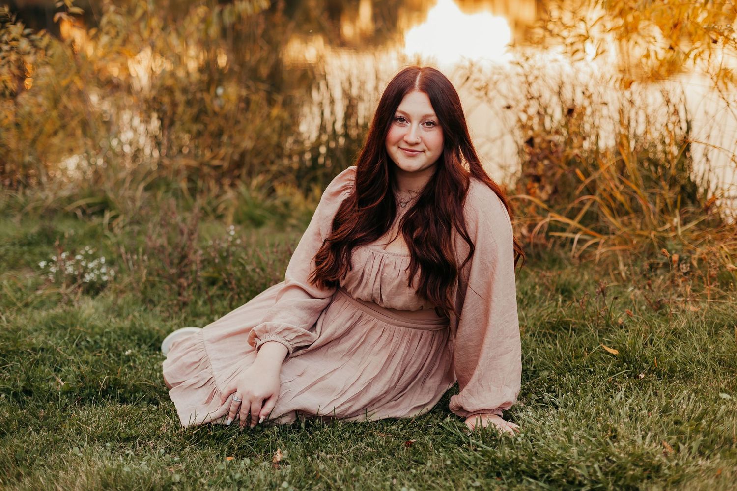 A senior girl sitting in the grass in front of a pond at  Minooka Park in Waukesha, Wisconsin wearing a knee-length pink dress.