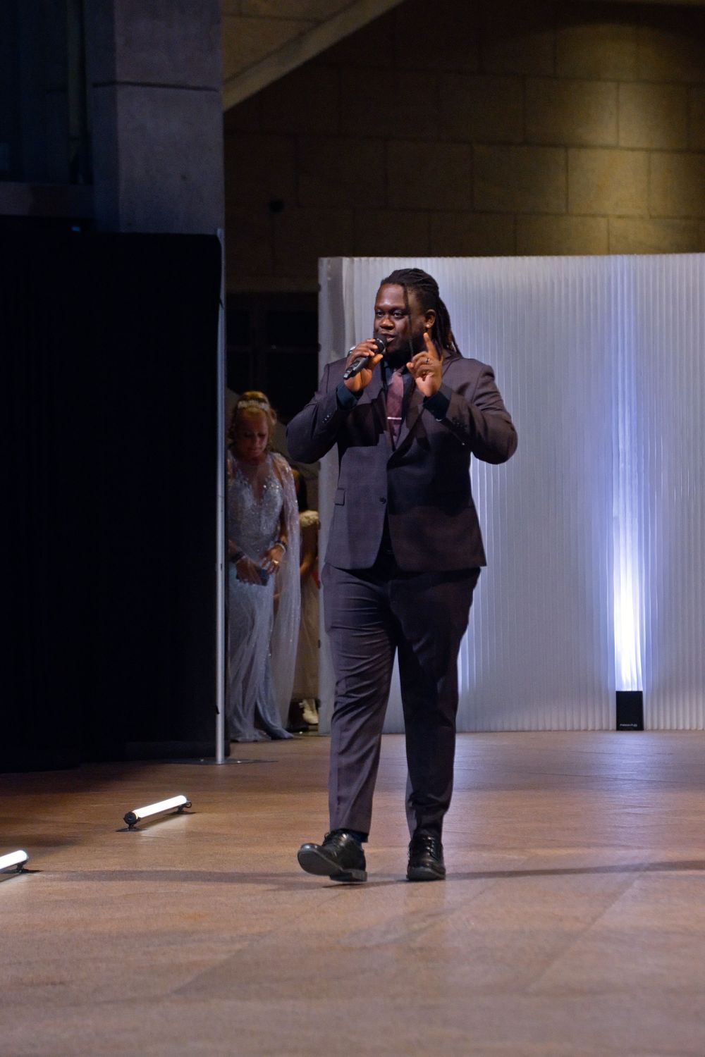A man in a dark suit speaks into a microphone on a runway stage during a fashion show event.