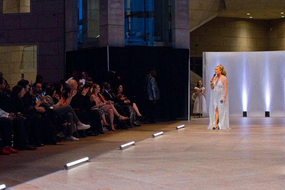 A model walks the runway in a white gown at a fashion show with seated audience members lining the catwalk.