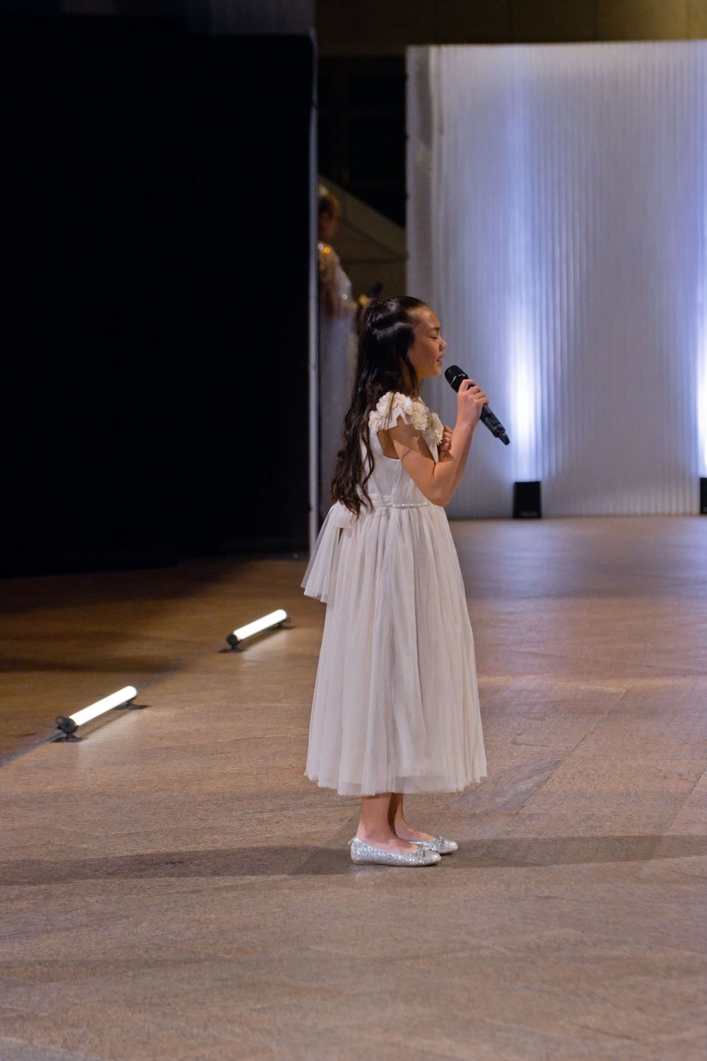 Young girl in white dress singing into microphone on stage at formal event.
