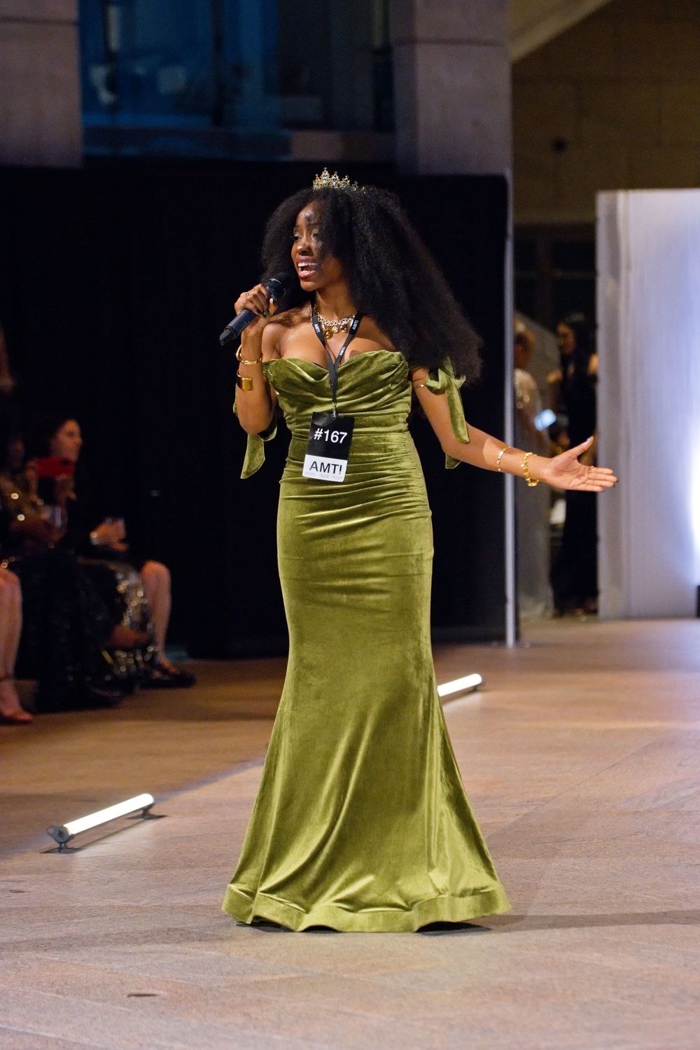Woman in olive green velvet gown wearing crown performs on runway holding microphone at formal pageant event.