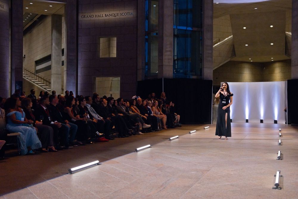 A model walks a lit runway at an indoor fashion show with an audience seated along the sides in a modern venue.