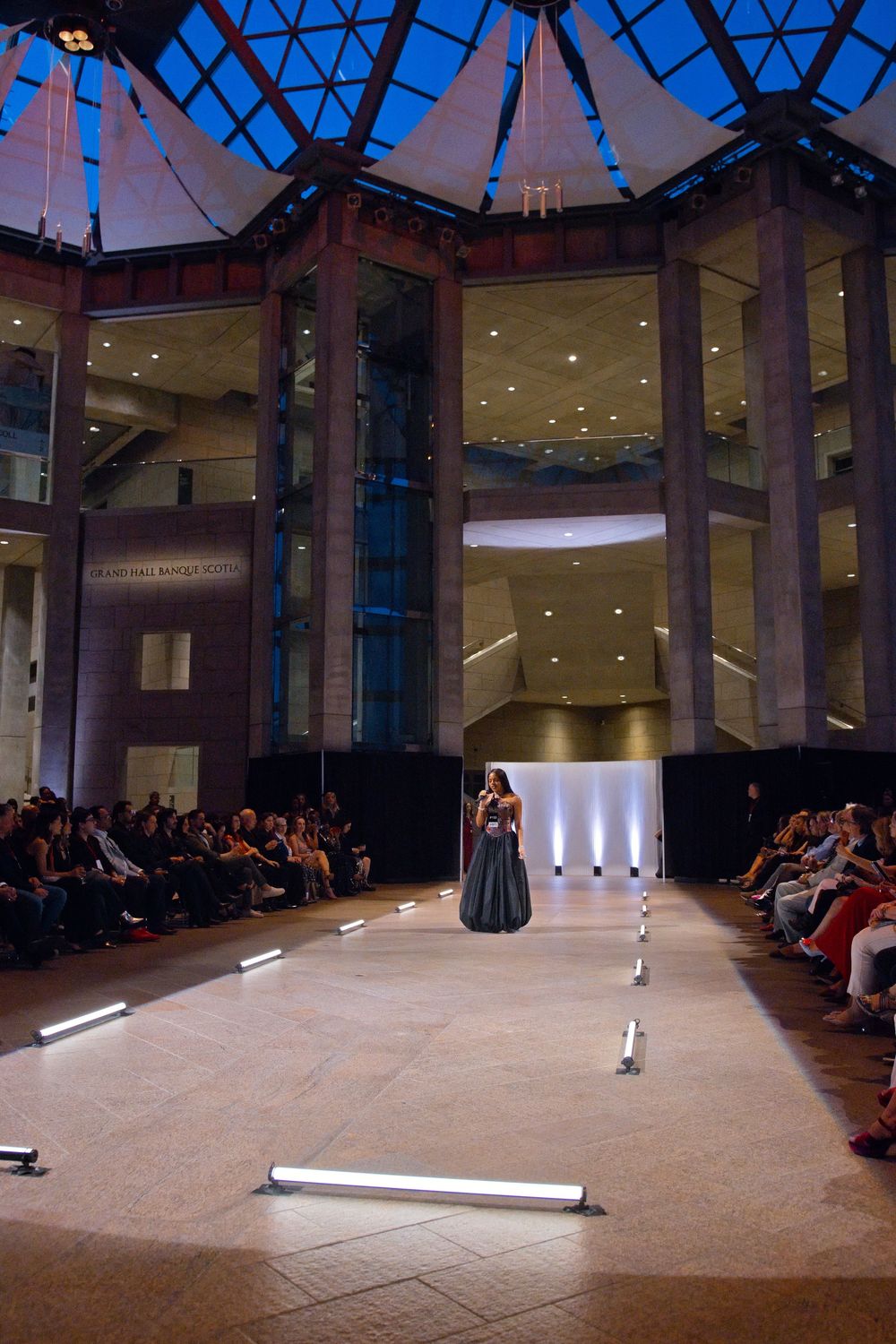 A woman in an elegant gown stands on a lit runway inside a grand domed venue during a fashion show.