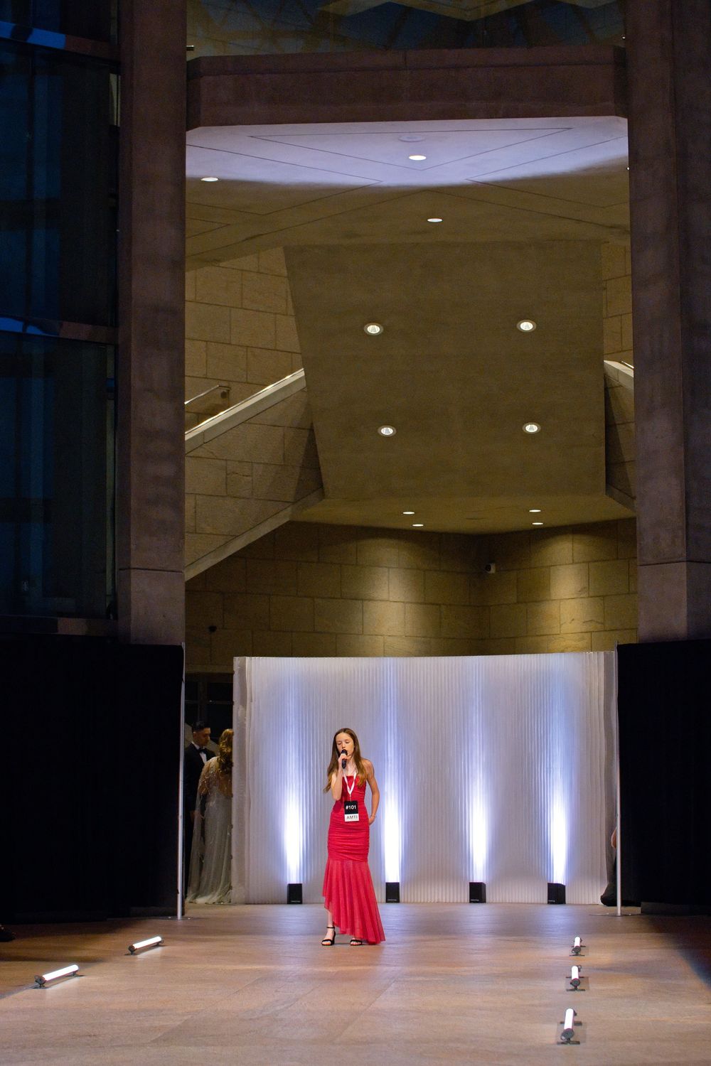 Woman in red gown performing on stage in elegant modern venue with dramatic lighting and architectural backdrop.