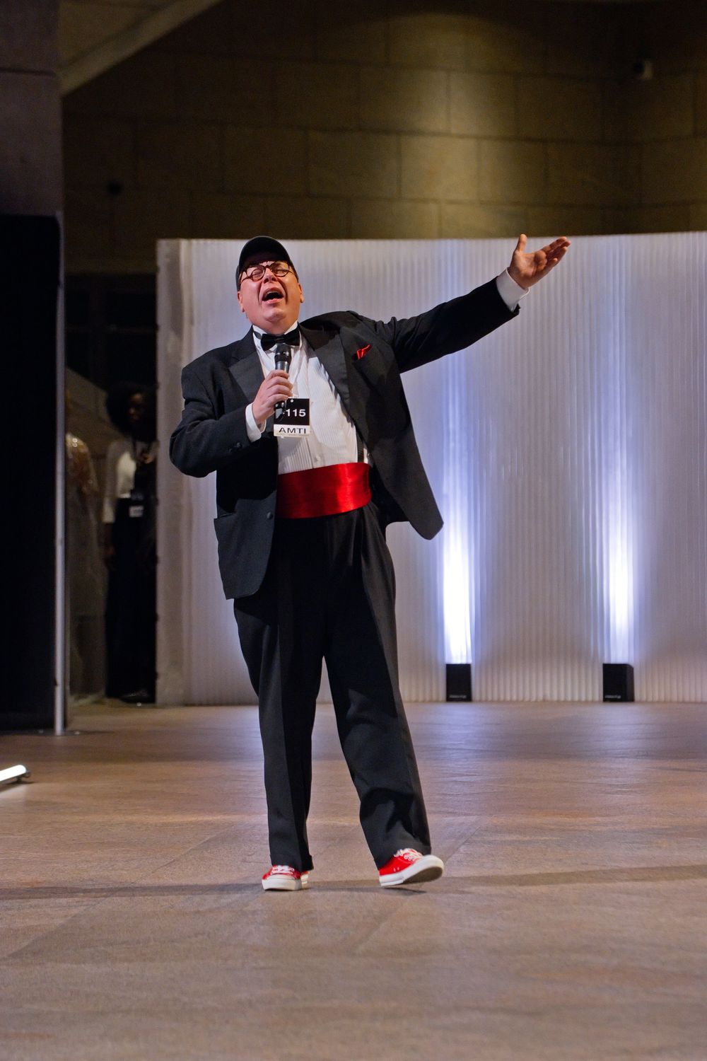 A performer in a black suit with red sash and sneakers sings energetically on a fashion show stage.