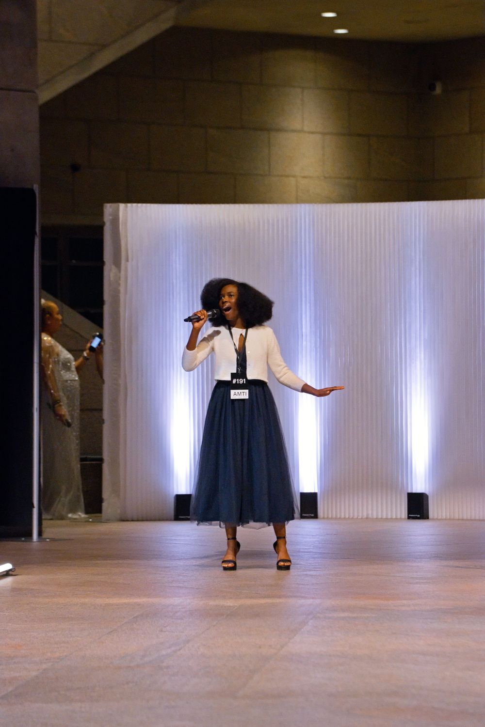 A woman in a navy tulle skirt and white blouse sings into a microphone on a lit stage.