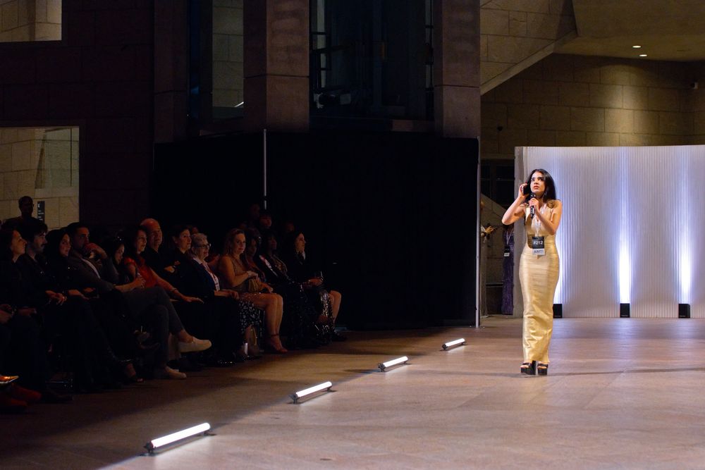 Woman in gold dress speaking on stage with microphone before seated audience in dimly lit venue.