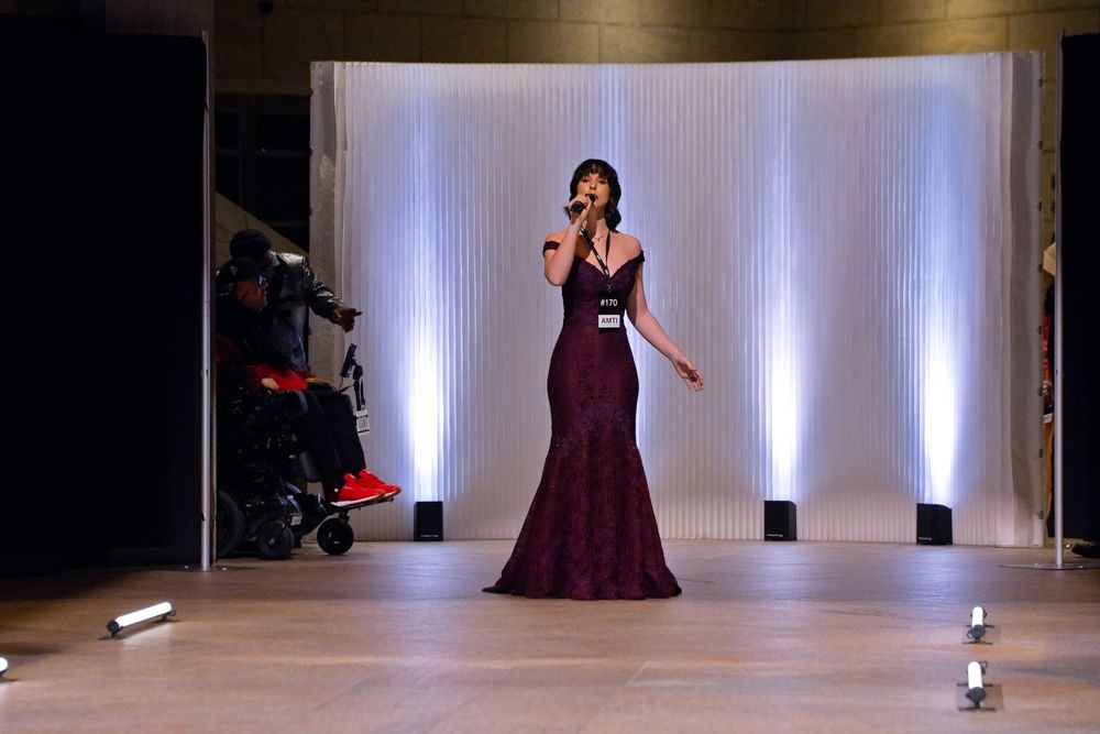 Woman in burgundy gown singing on stage at formal event with white backdrop lighting.