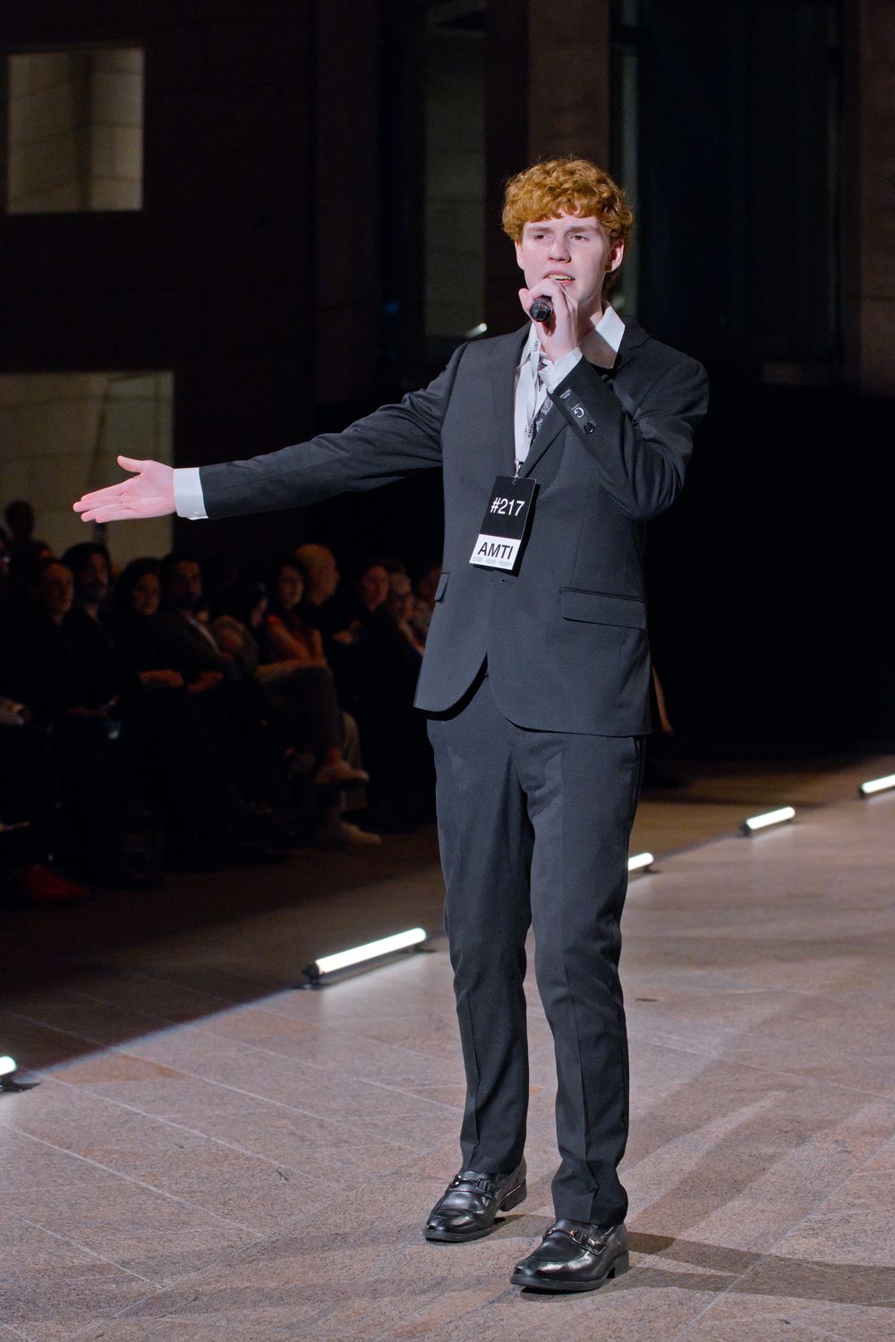 Young man with curly red hair in gray suit performing on runway stage with microphone and outstretched arm.