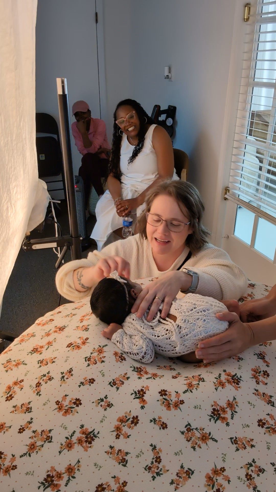 Woman in white sweater holds sleeping newborn baby while two women smile in background of a cozy room.