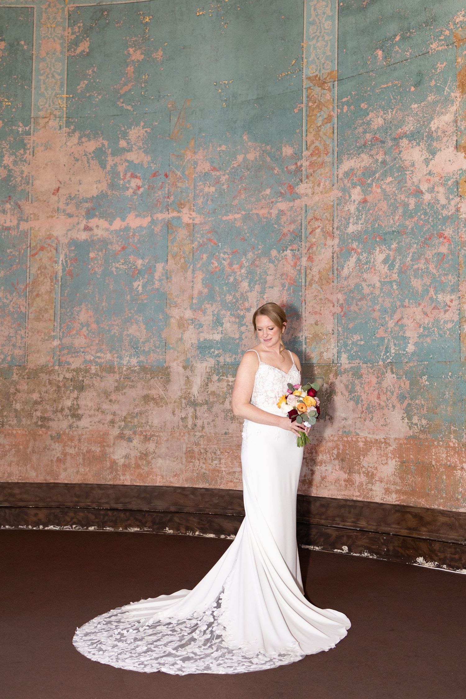 Bride in white satin mermaid gown with lace train holds colorful bouquet before ornate vintage wall in the Monastery Event Center.