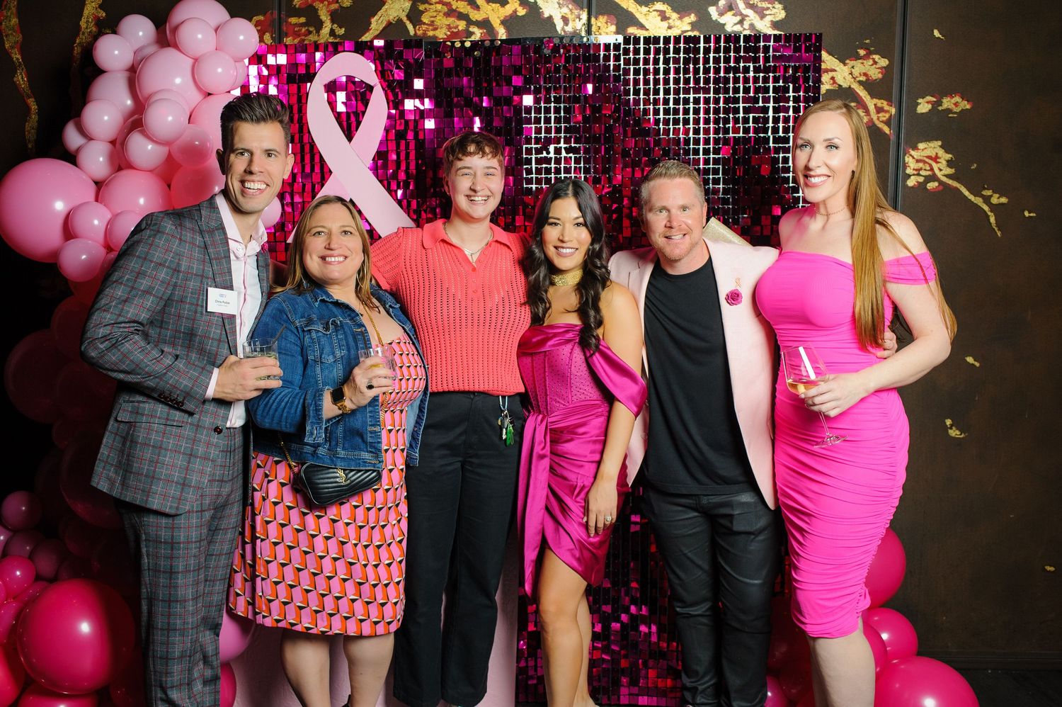 Group of six people posing at a pink breast cancer awareness event with balloon decorations and a sequin backdrop.