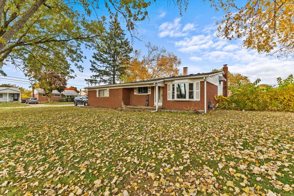 Brick ranch home with large front yard covered in fallen autumn leaves under blue sky.