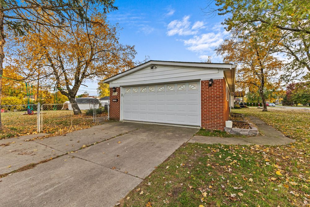Brick and vinyl detached garage with white door and concrete driveway surrounded by autumn trees.