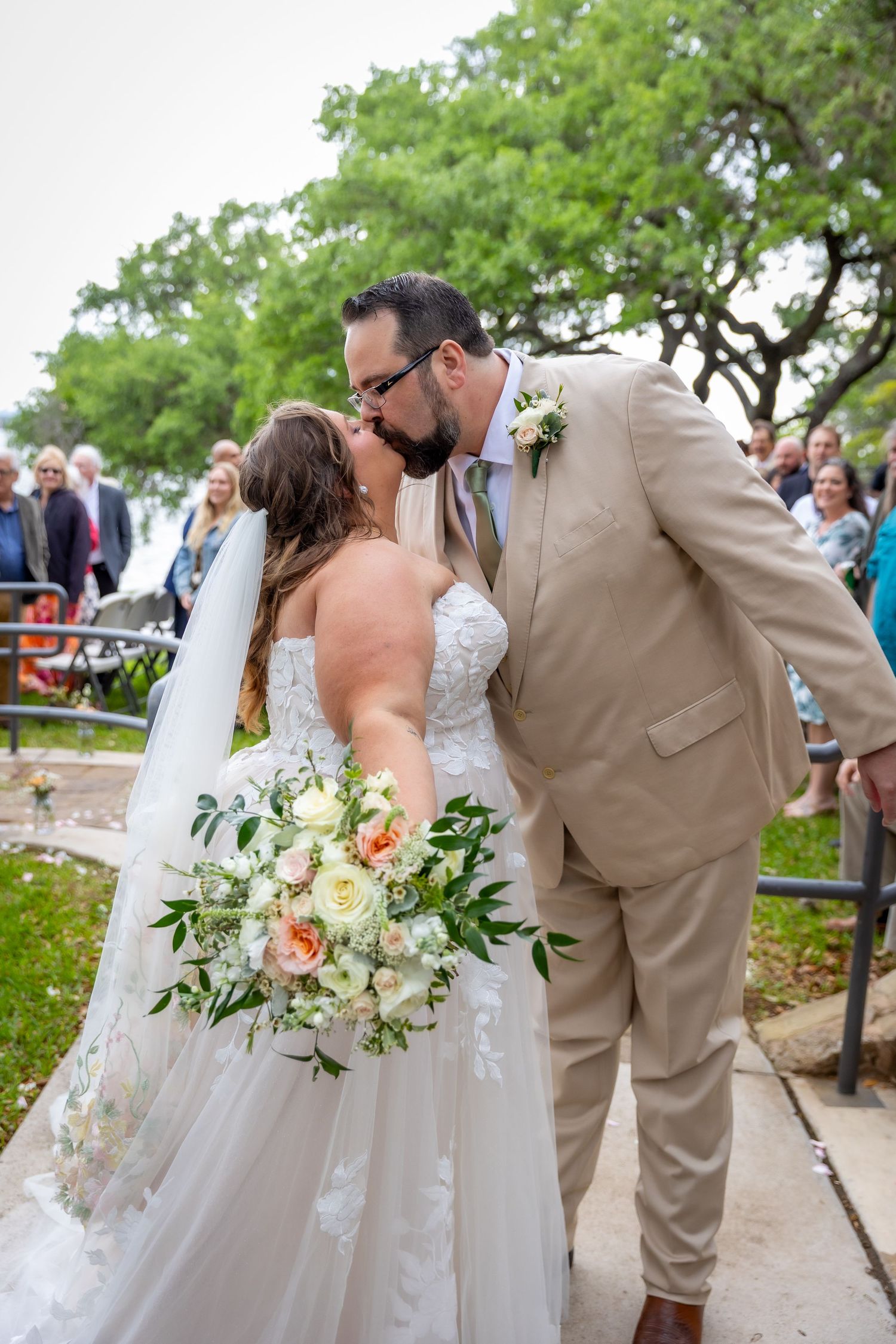 Bride and groom share first kiss at outdoor wedding ceremony, bride holding white and peach bouquet, groom in tan suit.