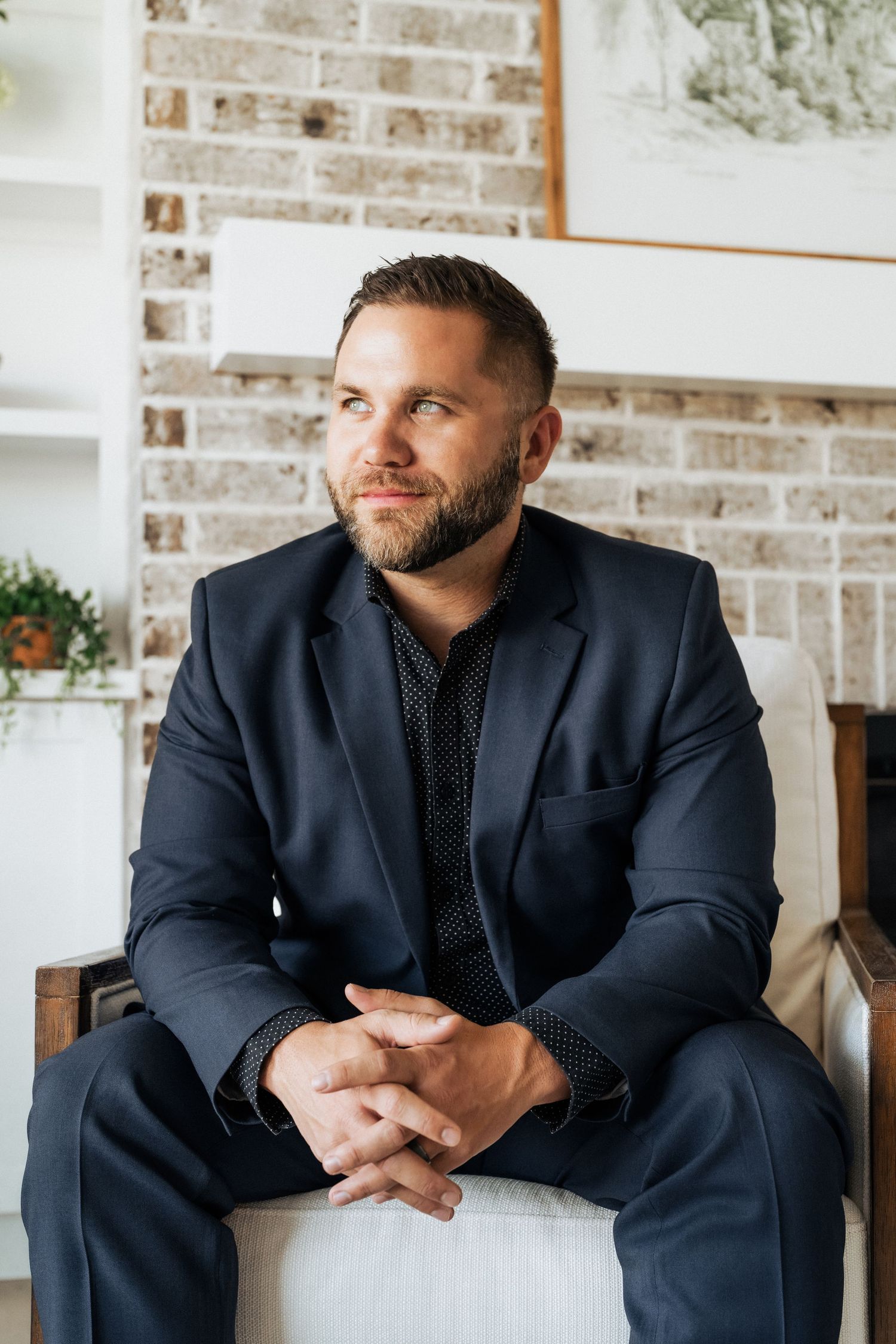 Man in navy suit sitting in chair with hands clasped, looking away thoughtfully against brick wall backdrop.