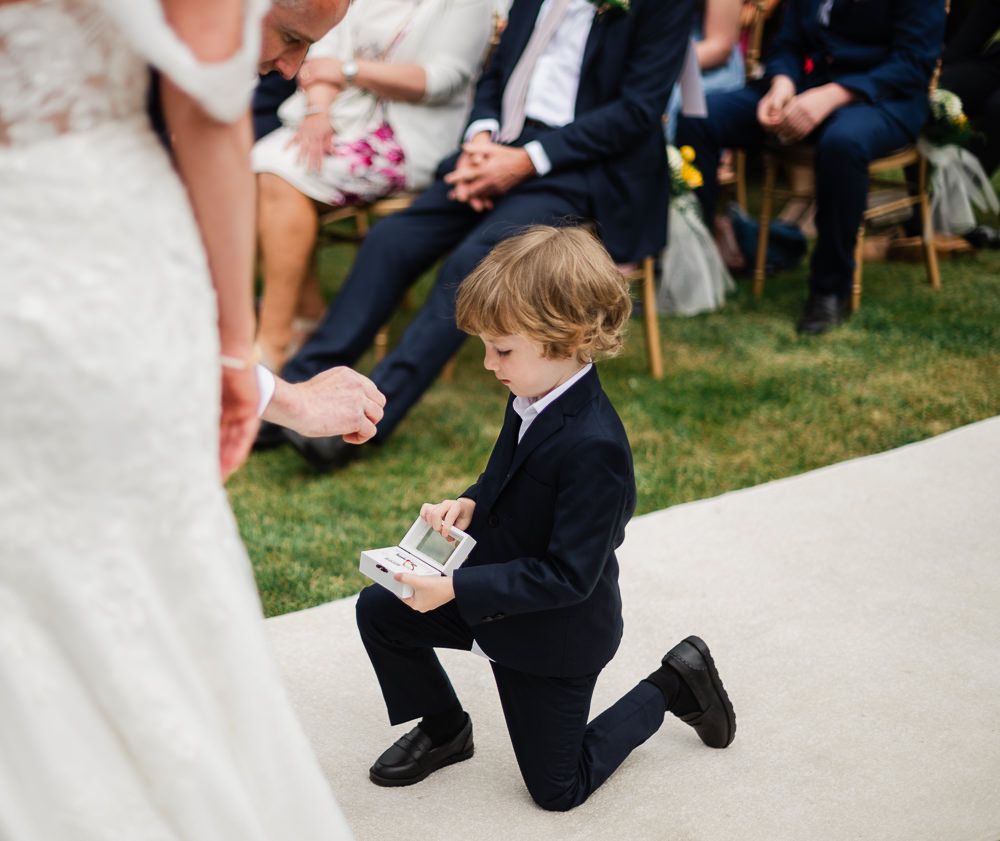 Young ring bearer in black suit kneeling on aisle, holding cards at outdoor wedding ceremony.
