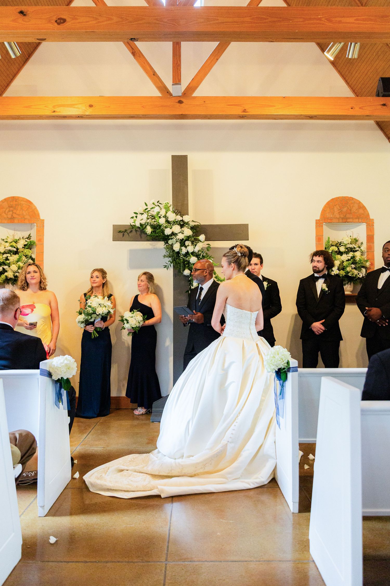 Bride in white gown walks down church aisle toward cross altar, flanked by bridesmaids in navy and groomsmen in tuxedos.