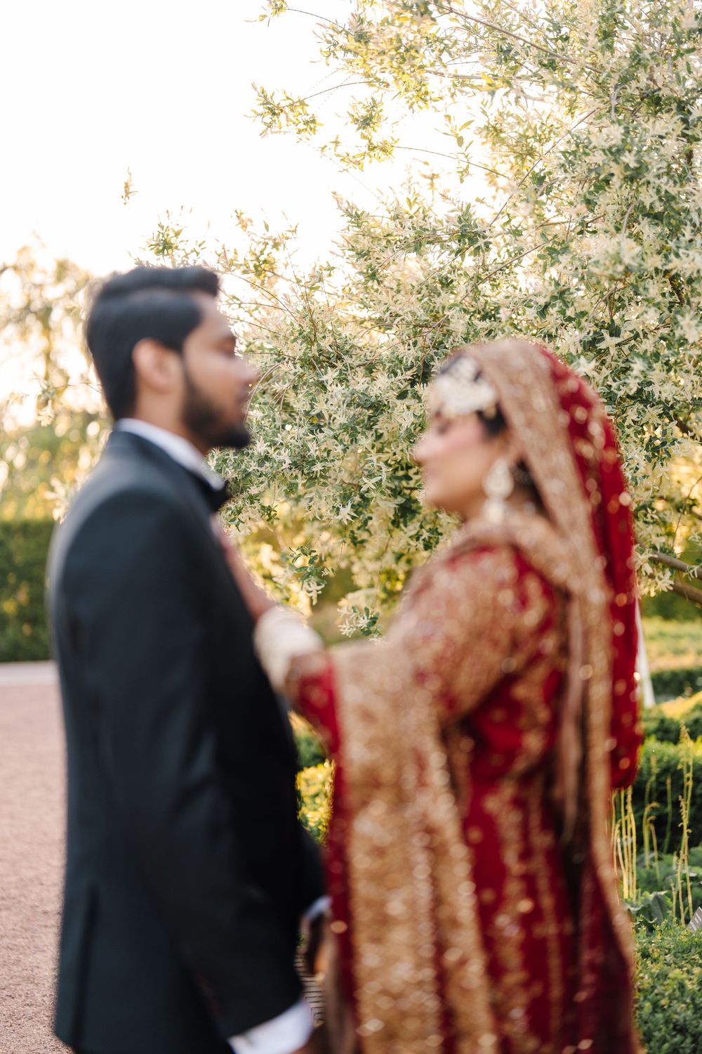 South Asian bride in red bridal lehenga and groom in black suit share a romantic moment outdoors.