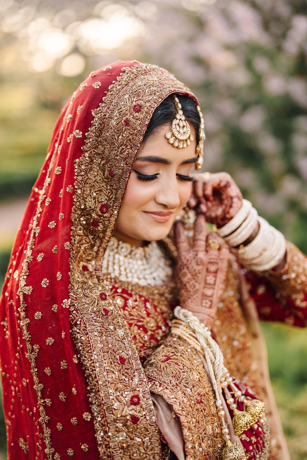 A bride in red and gold embroidered lehenga with mehndi hands, maang tikka, and dupatta, smiling softly outdoors.