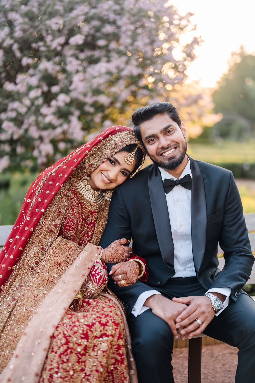 South Asian bride in red gold bridal lehenga and groom in black tuxedo posing together at outdoor wedding.