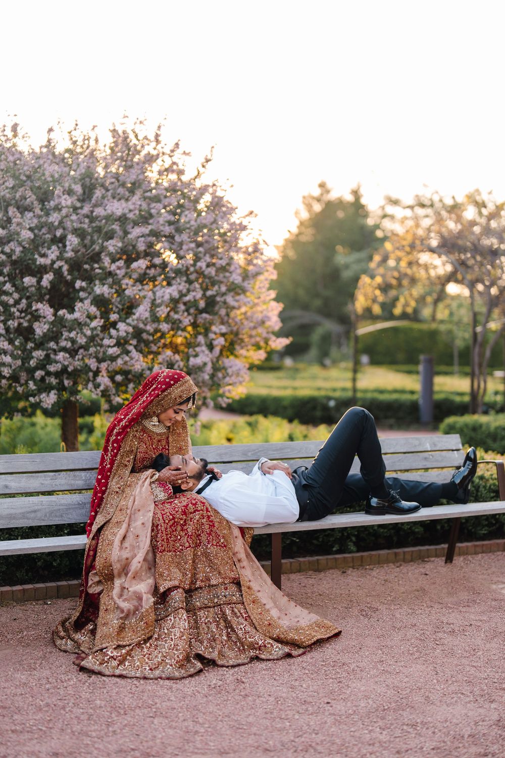 A bride in ornate golden red lehenga sits on a park bench with groom in white shirt resting beside her.