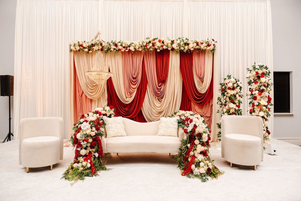 Elegant wedding stage with red and pink draped backdrop, floral arrangements, and white sofa seating.