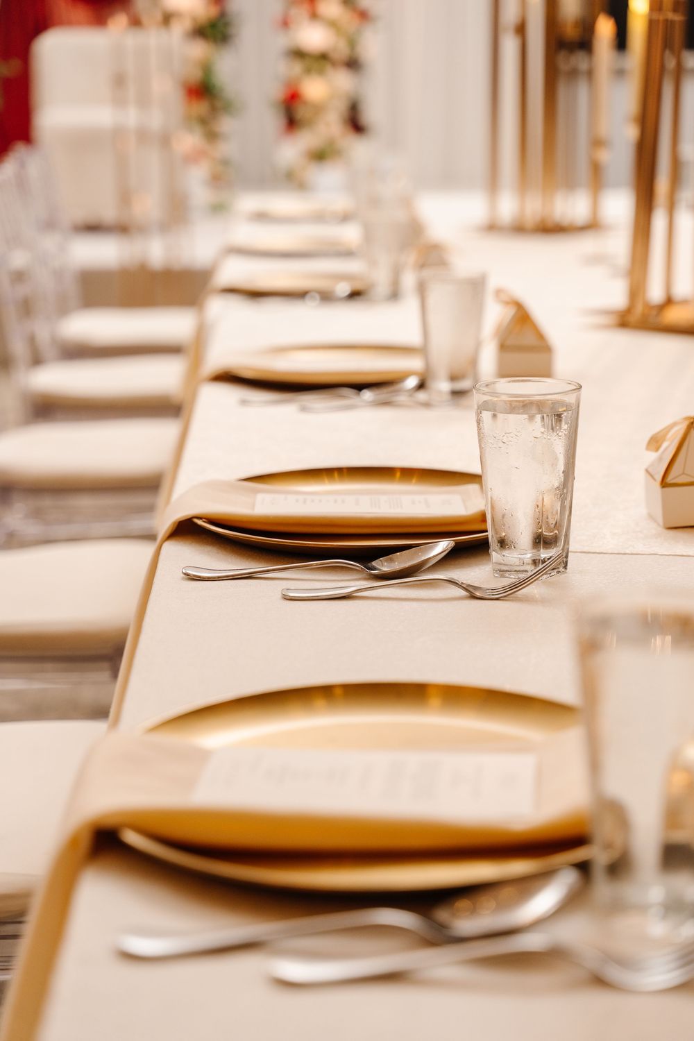 Elegant wedding reception table set with gold charger plates, silverware, menus, and crystal glasses on a white linen tablecloth.