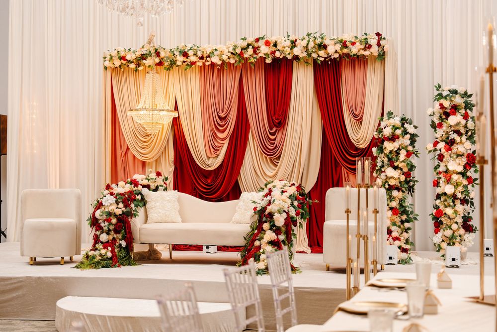 Elegant wedding stage with red and gold draped backdrop, white sofa, and floral arrangements of roses.