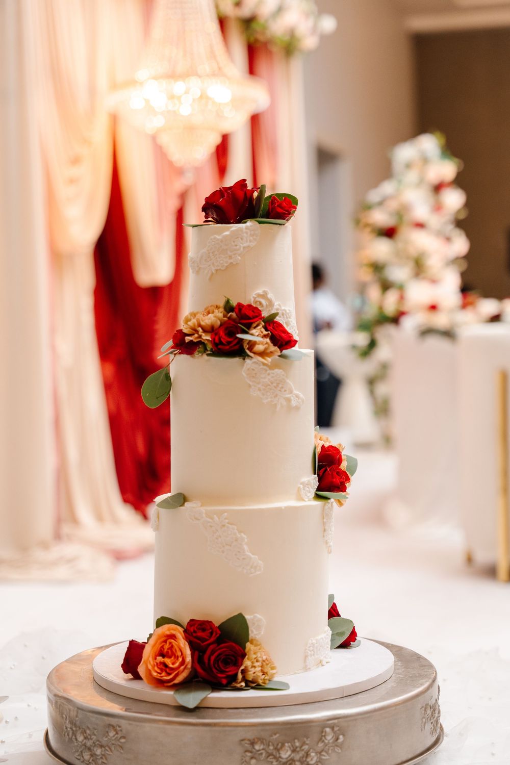 Elegant three-tier white wedding cake adorned with red roses on a silver stand at a reception.