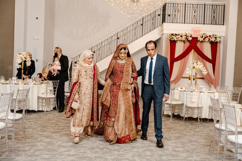 Bride in red bridal lehenga escorted by family members into a decorated wedding reception venue.