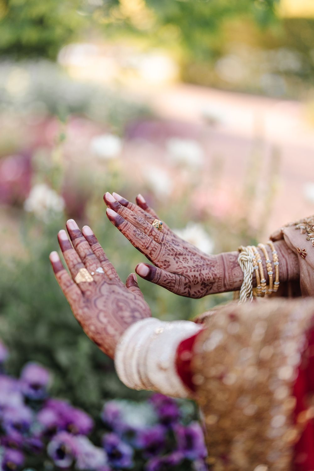 Close-up of Indian bride's hands adorned with intricate henna mehndi designs, gold bangles, and rings against garden backdrop.