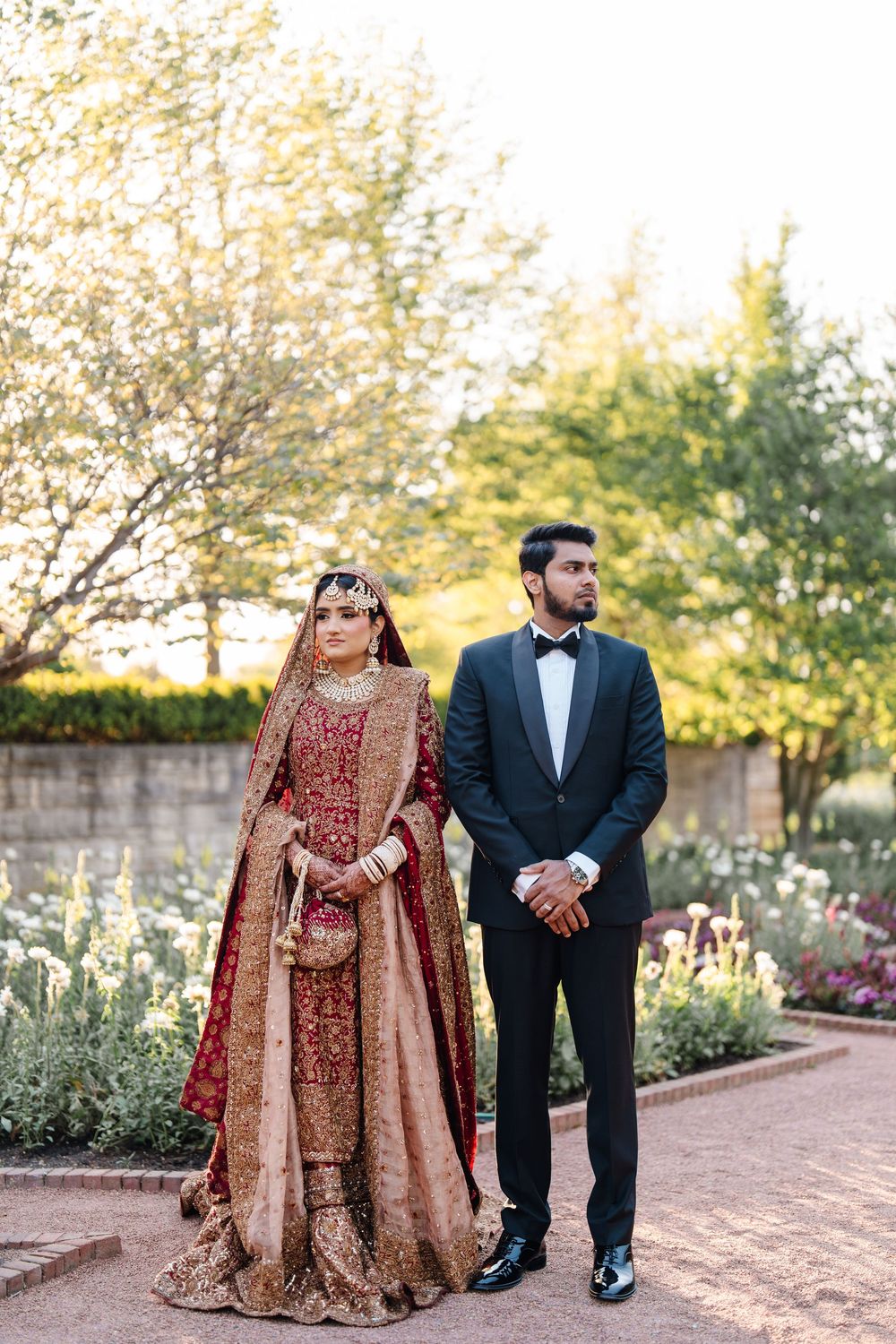 South Asian bride in gold and red lehenga stands with groom in black tuxedo in a garden wedding portrait.