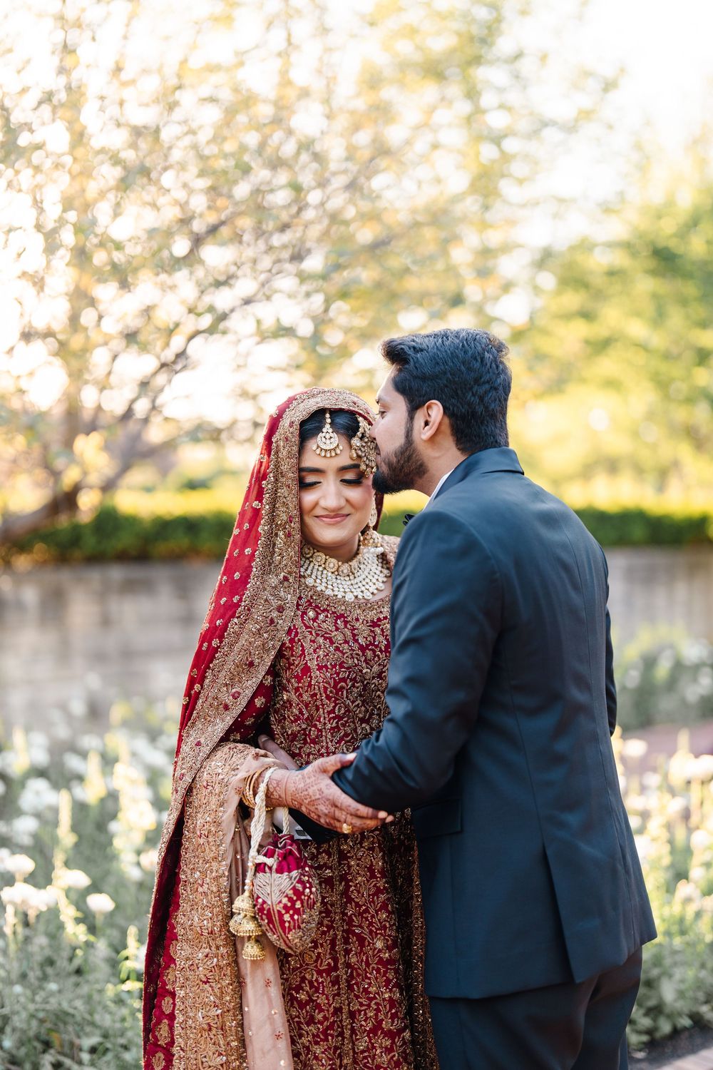A bride in red and gold bridal lehenga with groom in navy suit sharing a romantic moment in a garden.