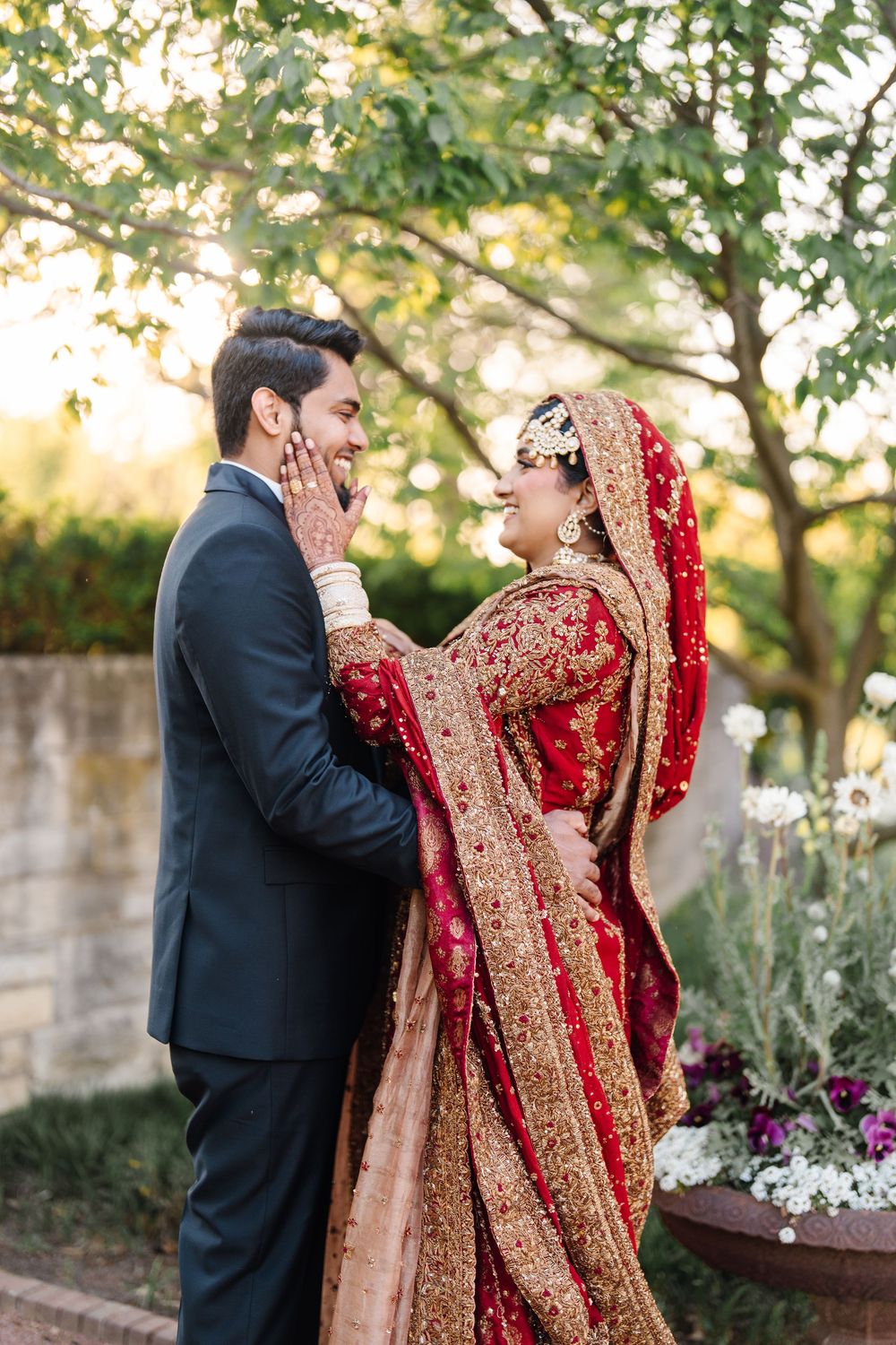 South Asian bride in red gold lehenga tenderly holds grooms face during romantic outdoor wedding portrait.