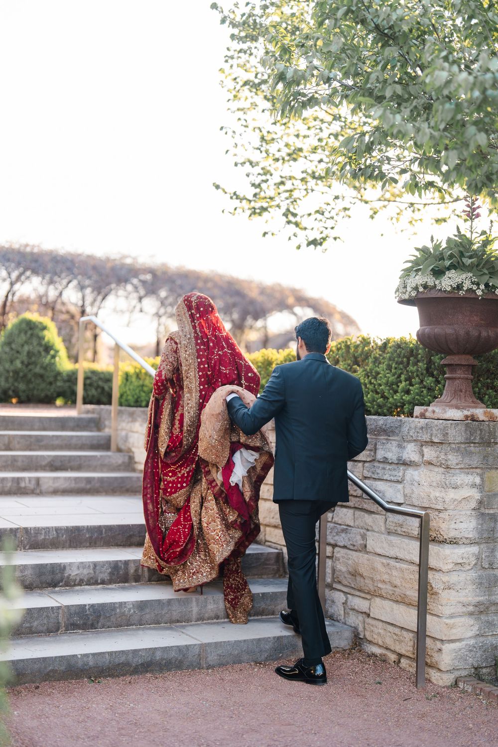Groom in dark suit assists bride in red embroidered lehenga up stone steps at outdoor wedding.