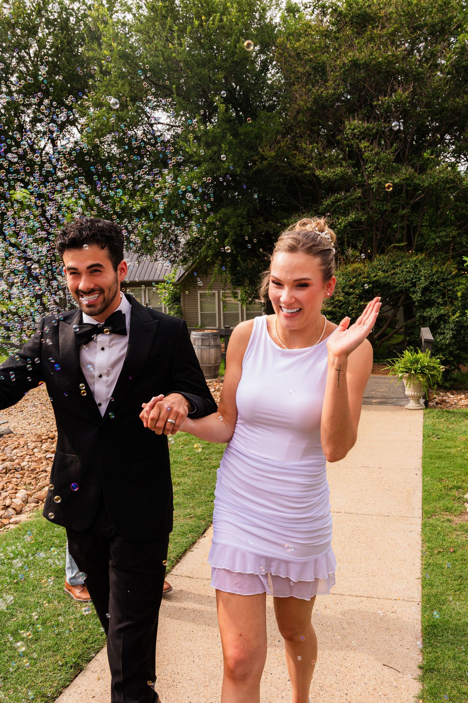 Happy couple in wedding attire walking outdoors, man in tuxedo and woman in white mini dress, surrounded by bubbles.